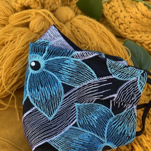 a closeup of the in full bloom mask, with embroidered flowers in teal, blue and white. with black and white embroidered leaves photographed on a yellow background with green leaves