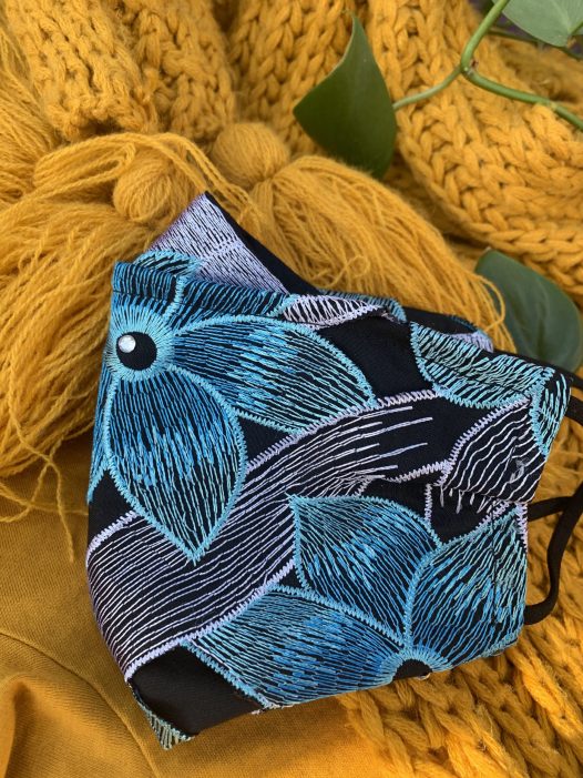 a closeup of the in full bloom mask, with embroidered flowers in teal, blue and white. with black and white embroidered leaves photographed on a yellow background with green leaves