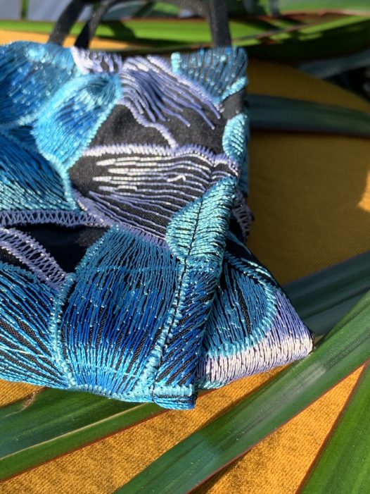 a closeup of the in full bloom mask, with embroidered flowers in teal, blue and white. with black and white embroidered leaves photographed on a yellow background with green leaves