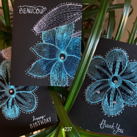 Three black greeting cards with leaves in the background. The cards feature a blue, teal and white embroidered flower, some with decorative gems, with words in white ink like “happy birthday” “thank you” and “merci beaucoup”